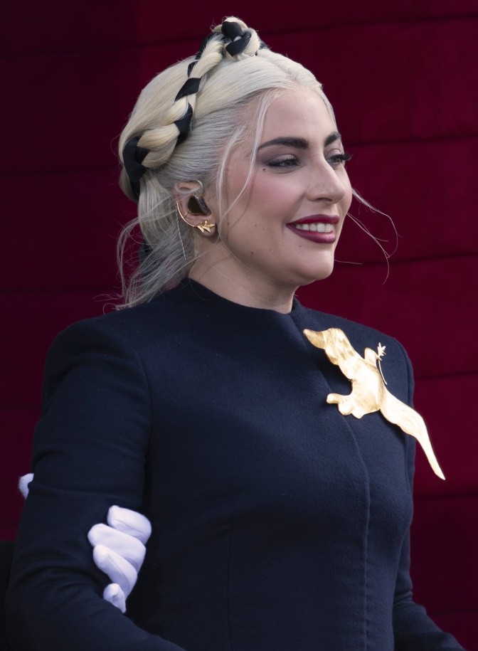 Lady Gaga mit einem schwarzen Band in die hellblonden Haare geflochten und in einem schwarzen  Kleid. Sie hat dunkelroten Lippenstift und lächelt. Am Revers hat sie eine goldene Friedenstaube als Brosche.