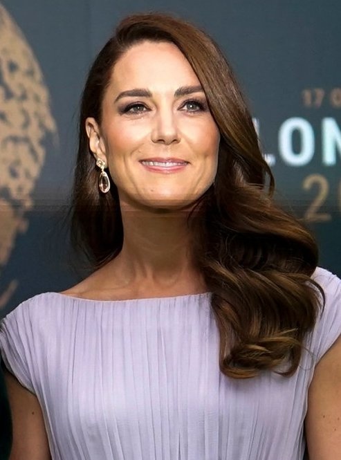Herzogin Kate mit gewellten, braunen Haaren in einem fliederfarbenen Kleid. Sie steht vor einer Logowand.
