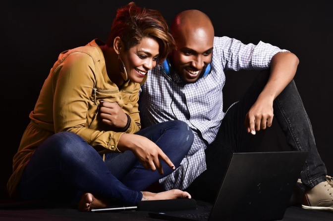 Ein dunkelhäutiges Paar sitzt auf dem Boden und schaut einen Film auf einem Tablet. Beide lachen, die Frau zeigt auf das Tablet.