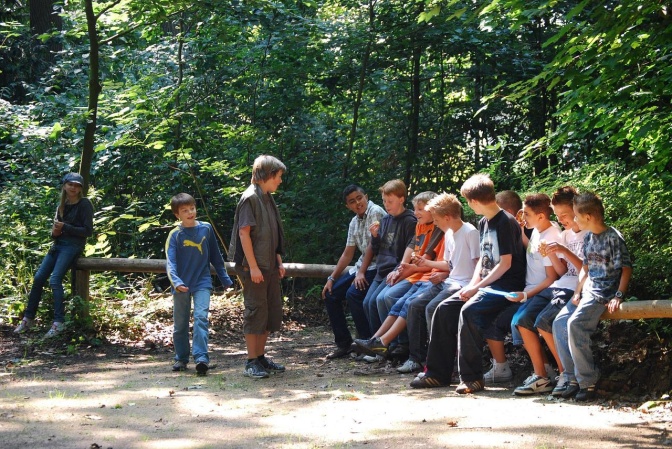 Eine Gruppe von Kindern und Jugendlichen im Wald. Sie sitzen auf einem Holzzaun und sprechen miteinander.