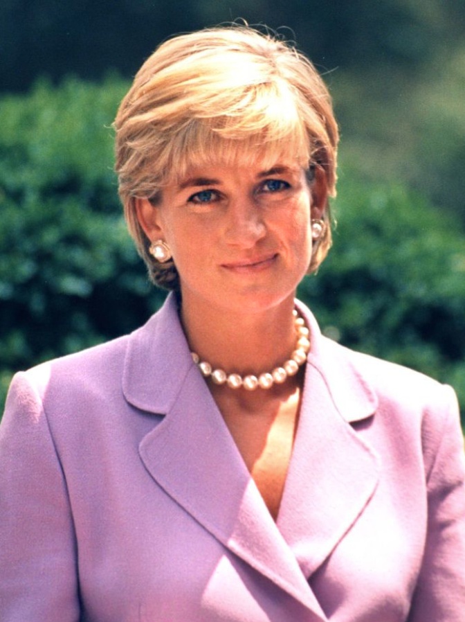 Prinzessin Diana in einem rosafarbenen Kostüm mit einer dicken Perlenkette. Sie hat kurze, blonde Haare und steht im Freien.