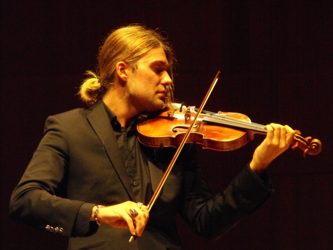 David Garrett mit einem blonden Pferdeschwanz im Anzug auf der Bühne. Er spielt Geige.