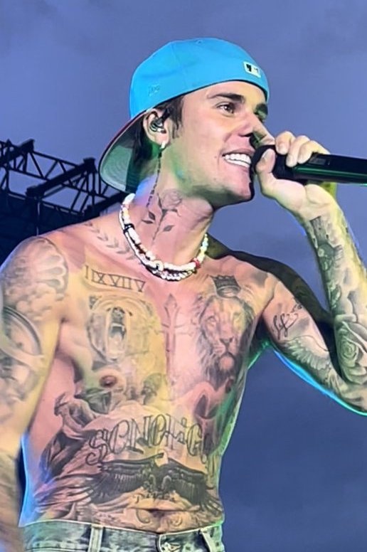 Justin Bieber mit einem freien, stark tätowierten Oberkörper. Er singt in ein Mikro und trägt eine verkehrtherum aufgesetzte Kappe in türkisblau.