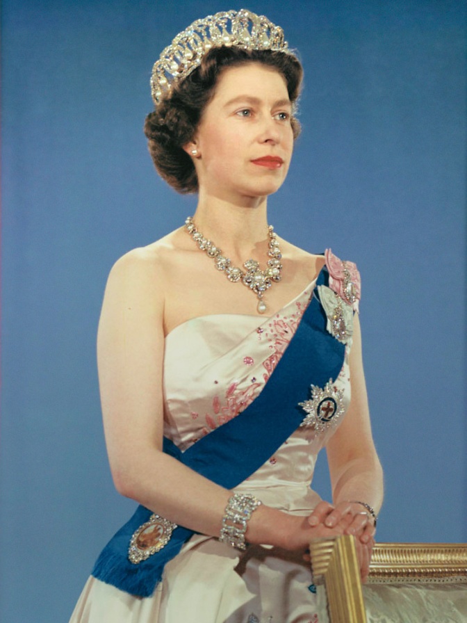 Die Queen in einem festlichen, hellen, schulterfreien Kleid mit einer blauen Schärpe. Sie trägt kostbaren Diamantschmuck und ein großes Diadem. An der Schärpe hat sie mehrere Orden angesteckt. Sie schaut ernst seitlich an der Kamera vorbei.