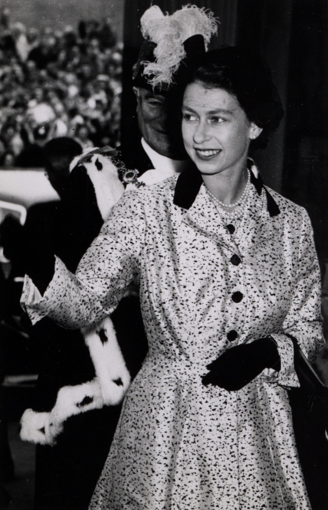 Schwarz-weiß Foto der Queen mit dunklen, in Wellen gelegten Haaren in einem getupften Mantel und schwarzen Handschuhen. Sie winkt.
