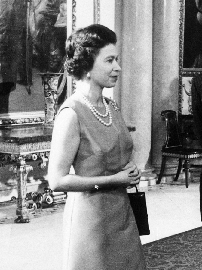 Schwarz-weiß Foto der Queen mit braunen, in Wellen gelegten Haaren in einem schlichten Etuikleid mit doppelreihiger Perlenkette.