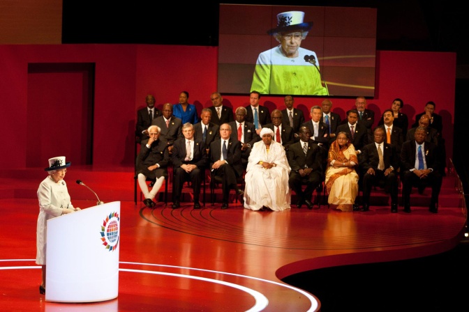 Die Queen steht in Kostüm und mit Hut an einem Rednerpult. Politiker*innen verschiedener Staaten sitzen unter einem großen Bildschirm, auf dem die Rede der Queen übertragen wird.