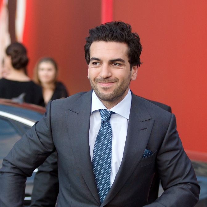 Elyas M'Barek in einem grauen Anzug mit Krawatte. Er ist gerade aus einem Auto ausgestiegenen lächelt.