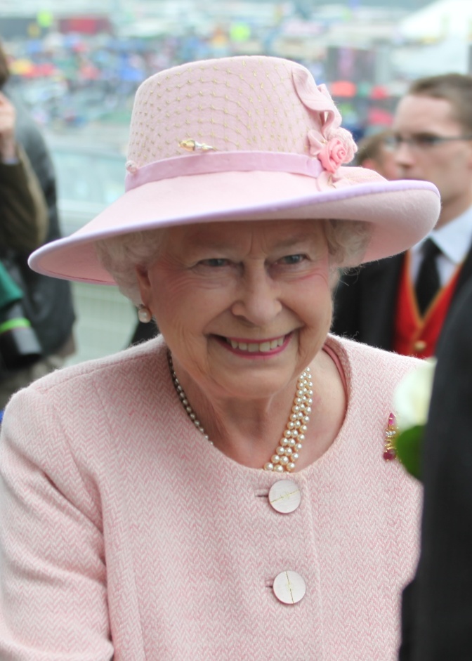 Die Queen in einem rosafarbenen Kostüm mit passendem Hut und Perlenkette. Sie lächelt. Um sie herum Menschen.