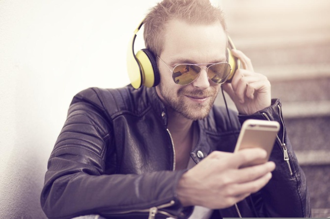 Ein Mann mit Vollbart und Sonnenbrille hat Kopfhörer auf und ein Handy in der Hand.