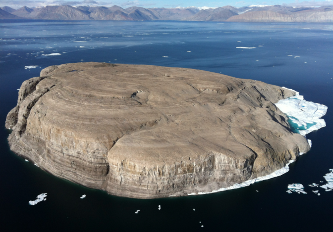 Luftaufnahme einer sehr kleinen, kargen Insel aus grauem Gestein. Sie ist vollkommen leer. Im Meer darum herum Schwimmen Eisbrocken.