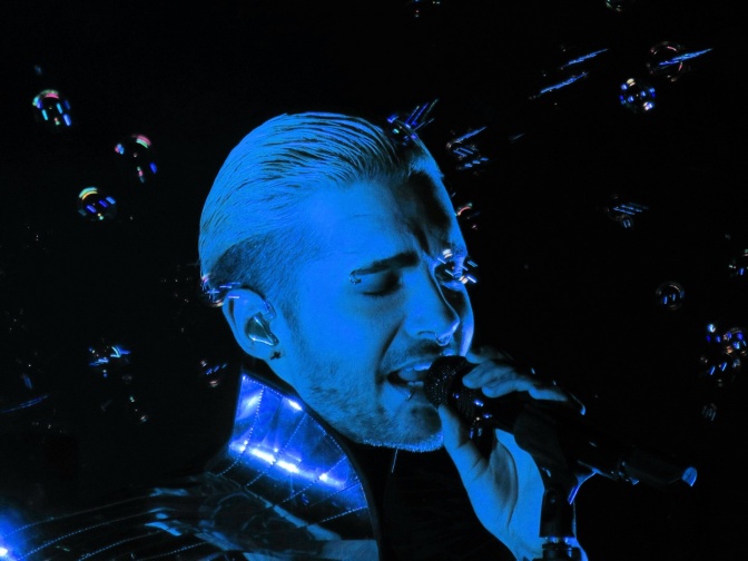 Ein blau eigefärbtes Foto von Bill Kaulitz auf der Bühne. Er singt in ein Mikro.