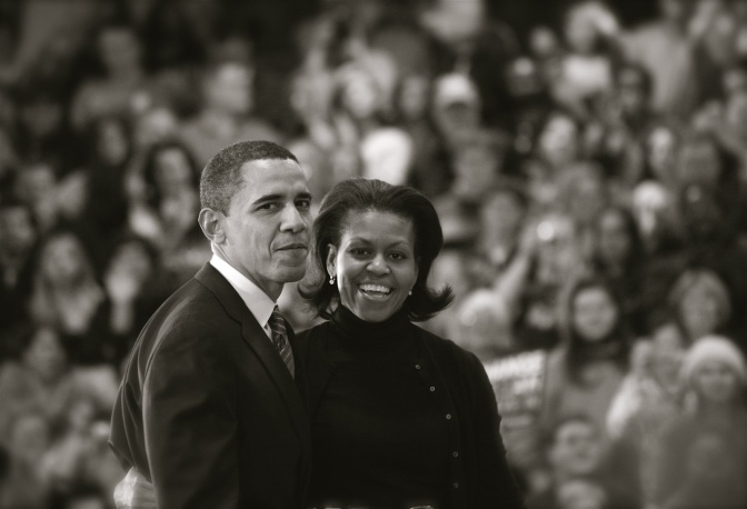 Schwarz-weiß-Foto von Barack und Michelle Obama vor einer großen Menschenmenge. Beide stehen Arm in Arm und lächeln.