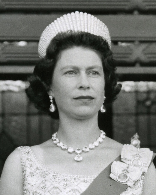 Schwarz-weiß-Foto der Queen mit Perlenkette und einem kleinen weißen Hut. Sie trägt ein Kleid und darüber eine mit Orden verzierte Schärpe.