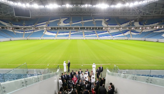 Eine Gruppe von Männern steht am Eingang der Rasenfläche eines Fußballstadions. Mehrere Menschen werden interviewt und gefilmt. Einige der Männer tragen klassische arabische Kleidung.