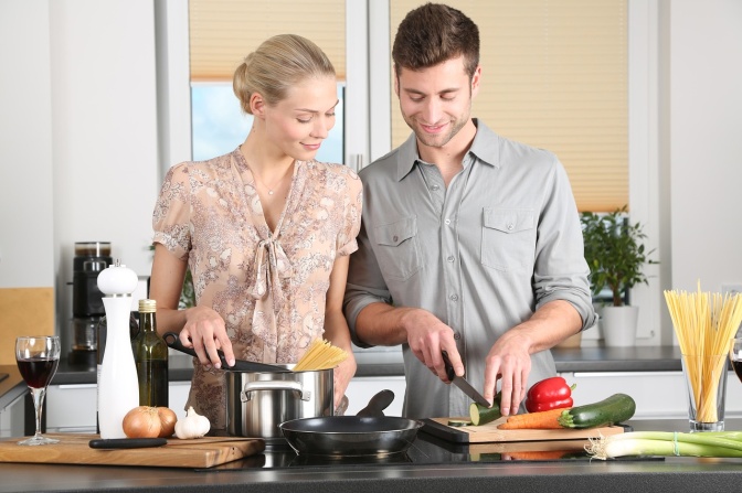 Ein Mann und eine Frau stehen zusammen in der Küche. Er schneidet Gemüse, sie sieht ihm bewundernd lächelnd dabei zu.