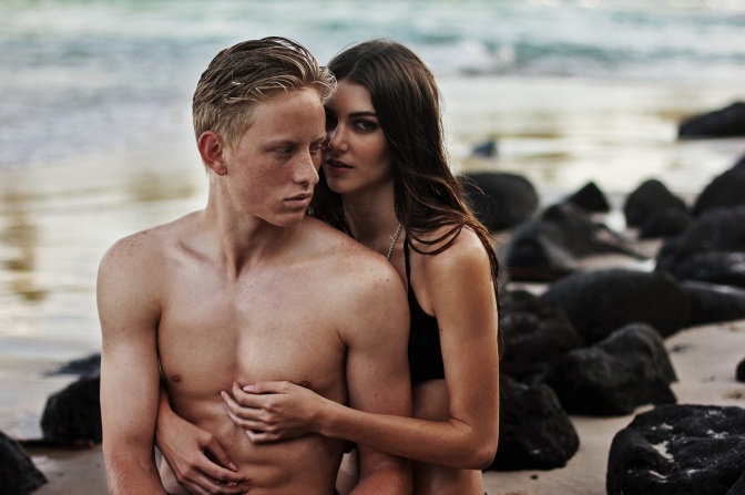 Eine Frau im Bikini steht hinter einem Mann mit nacktem Oberkörper und umarmt ihn. Beide sind am Strand.
