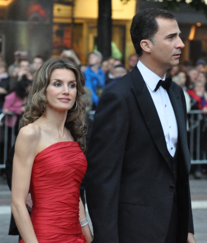 Felipe und Letizia in festlicher Kleidung. Sie trägt ein schulterfreies rotes Kleid, er Anzug und Krawatte. Sie halten sich an den Händen.