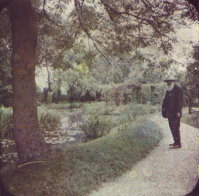 Claude Monet an einem Seerosenteich neben einem Baum. Er trägt Anzug und Hut und hat einen langen, weißen Bart.