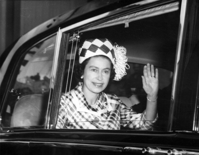 Schwarz-weiß-Foto der Queen winkend in einem schwarzen Auto. Sie trägt einen karierten Mantel mit kariertem Hut.