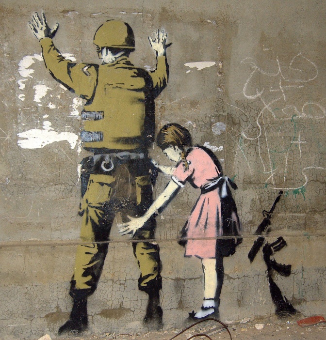 Ein Soldat steht mit ausgestreckten Armen an eine Hauswand gelehnt und wird von einem kleinen Mädchen in rosa Kleid abgetastet.