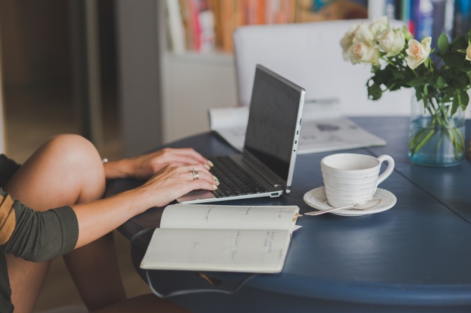 Eine Frau sitzt am Tisch und arbeitet am Laptop. Neben dem Laptop steht eine Tasse Kaffee.