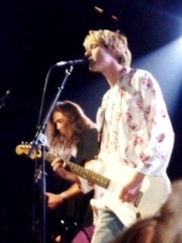 Kurt Cobain steht mit Gitarre am Mikro, andere Mitglieder der Band Nirvana sieht man verschwommen im Hintergrund.