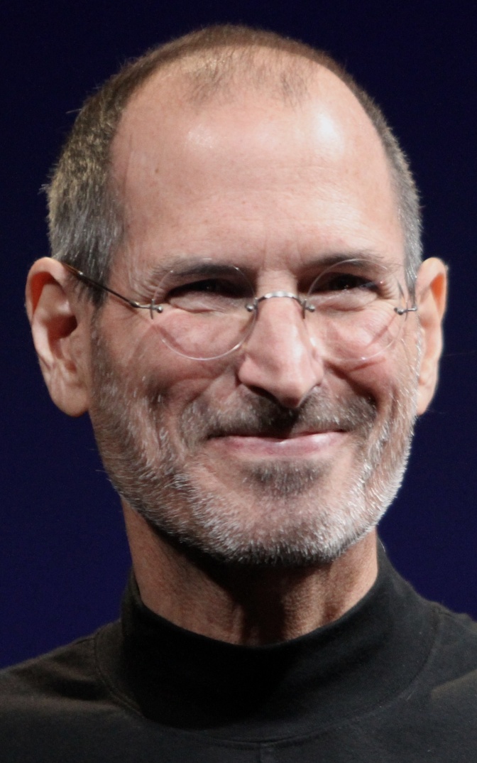 Steve Jobs mit Stirnglatze und einer kleinen, runden Brille. Er trägt ein schwarzes T-Shirt und lächelt in die Kamera.