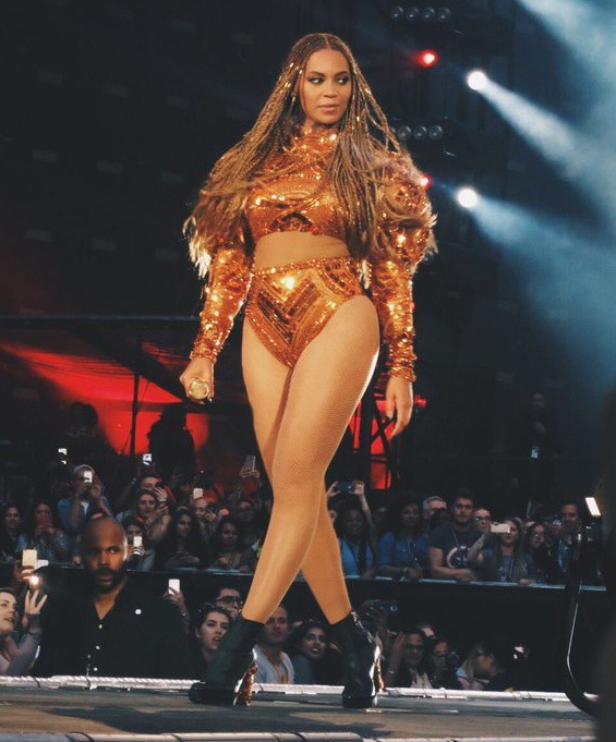 Beyoncé in einem bromzefarbenen Bühnenkostüm mit langen, geflochtenen Köpfchen. Sie wird von Scheinwerfern angestrahlt und schaut ins Publikum.