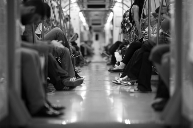 Schwarz-weiß-Foto von Menschen, die sich in 2 Reihen in einer Bahn gegenüber sitzen. Manche lesen oder sind mit dem Handy beschäftigt.