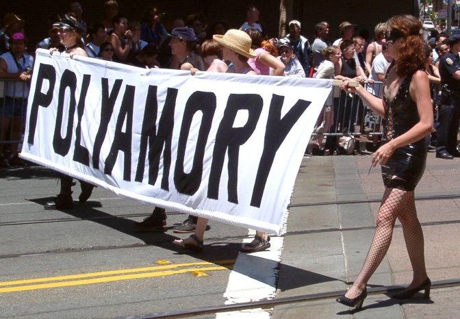 Menschen laufen bei einer Demonstration mit einem großen Schild mit dem Begriff Polyamory über die Straße.