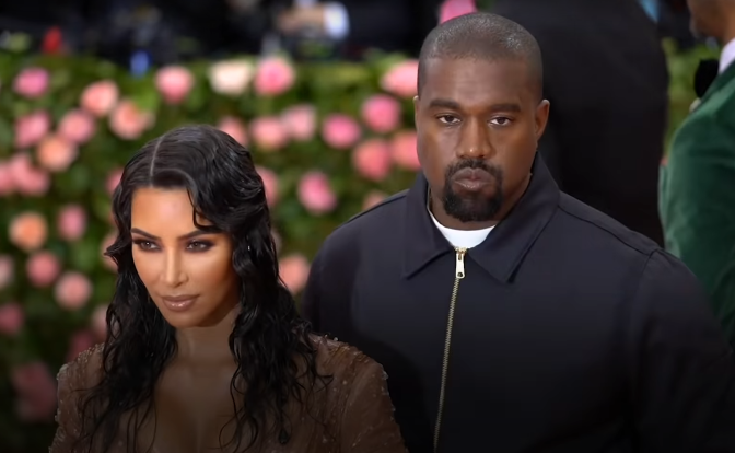 Kanye West und Kim Kardashian in dunkler Kleidung bei einem öffentlichen Auftritt. Im Hintergrund eine blühende Hecke und weitere Menschen.