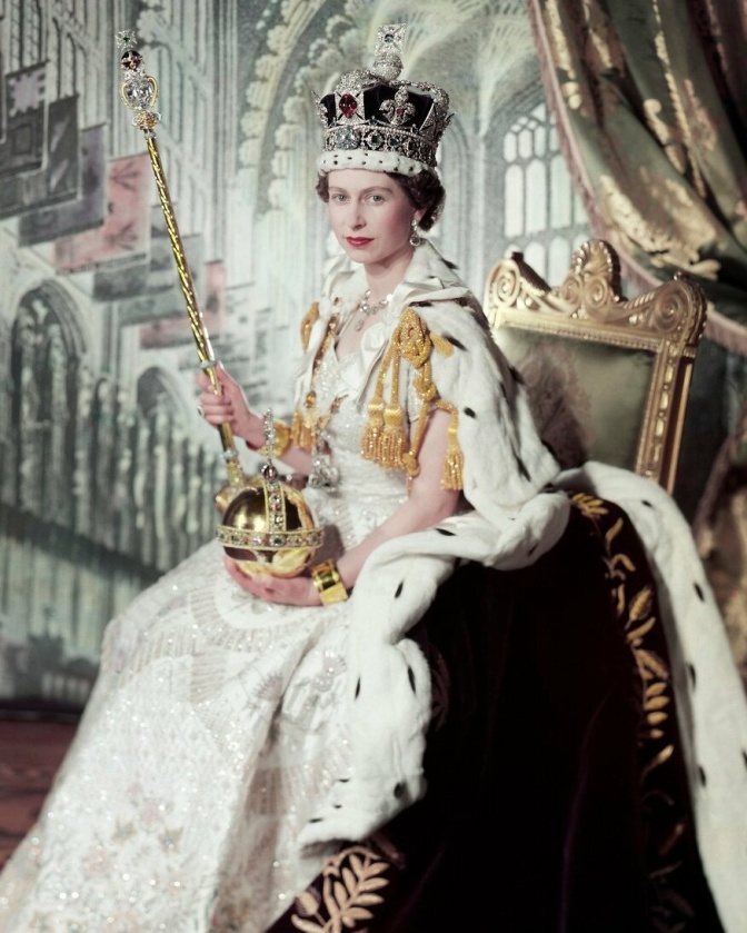 Die Queen in einem festlichen Kleid und mit einem Hermelinmantel. Sie trägt eine Krone und sitzt auf dem Thron.