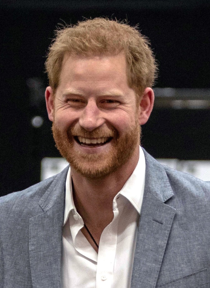 Prinz Harry mit roten Haaren und Dreitagebart. Er lächelt übers ganze Gesicht. Er trägt ein weißes Hemd und ein graues Sakko.