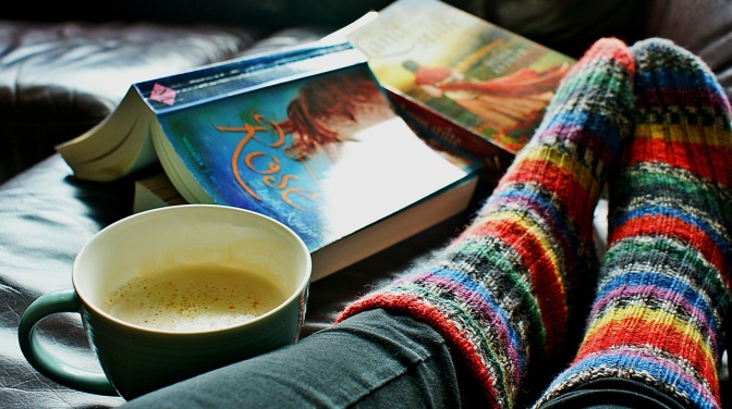 Eine Person mit Wollsocken sitzt auf einer Couch, Daneben  eine Tasse Kaffee und ein aufgeschlagenes Buch
