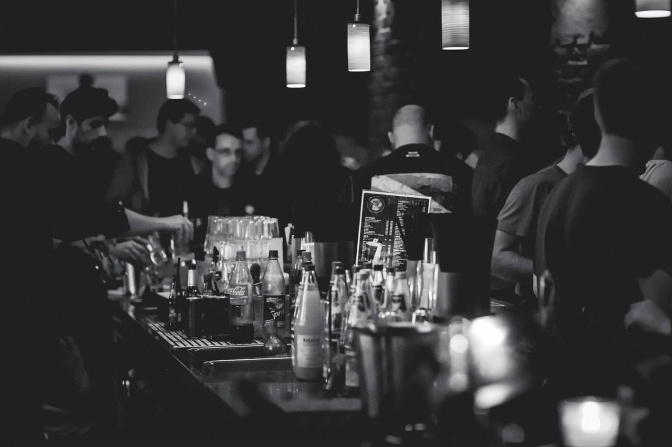 Schwarz-weiß-Foto von Menschen in einer Bar. Auf dem Tresen stehen Flaschen.