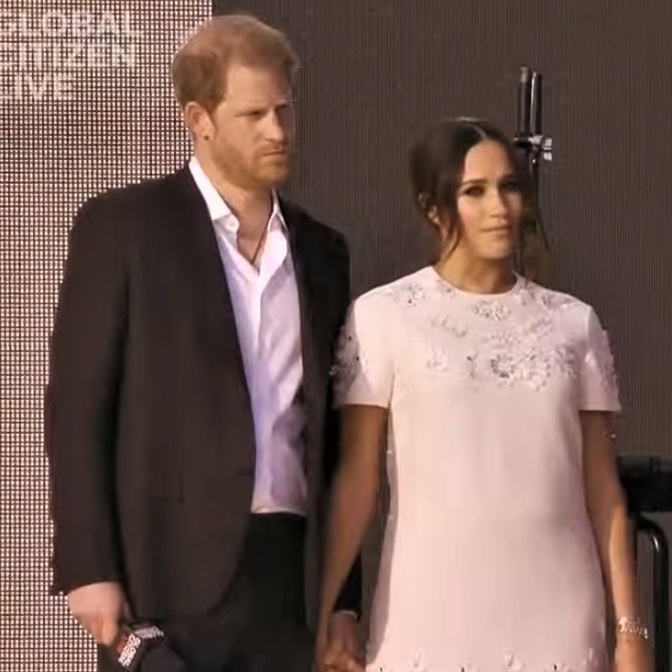 Prinz Harry und Herzogin Meghan stehen in festlicher Kleidung auf der Bühne und halten sich an den Händen.