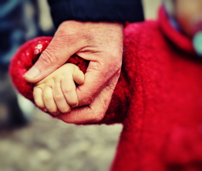 Die Hand einer alten Person hält die Hand eines Kleinkinds in rotem Mantel.