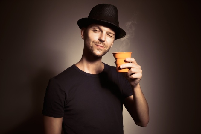 Ein Mann in schwarzem T-Shirt und mit schwarzem Hut hat einen Kaffeebecher in der Hand.