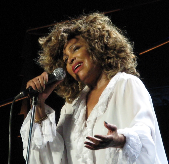 Tina Turner singt in ein Mikro und gestikuliert mit der Hand. Sie trägt eine Perücke und ein weißes Oversize Hemd.