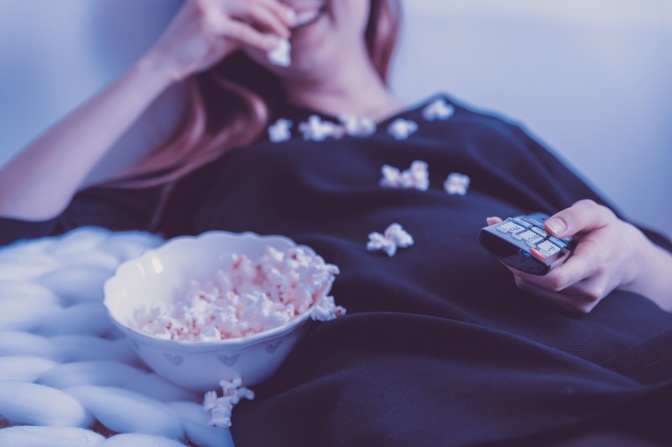 Eine Frau liegt mit der Fernbedienung in der Hand auf der Couch. Sie isst Popcorn und ihr Shirt ist mit Popcorn vollgekrümelt.