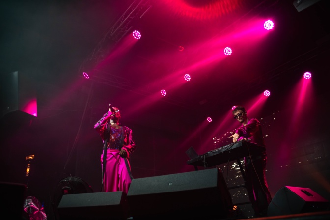 Musiker*innen auf einer dunklen, nur von lilafarbenen Strahlern beleuchteten Bühne. Ein Sänger und ein Keyboarder sind schemenhaft zu sehen.