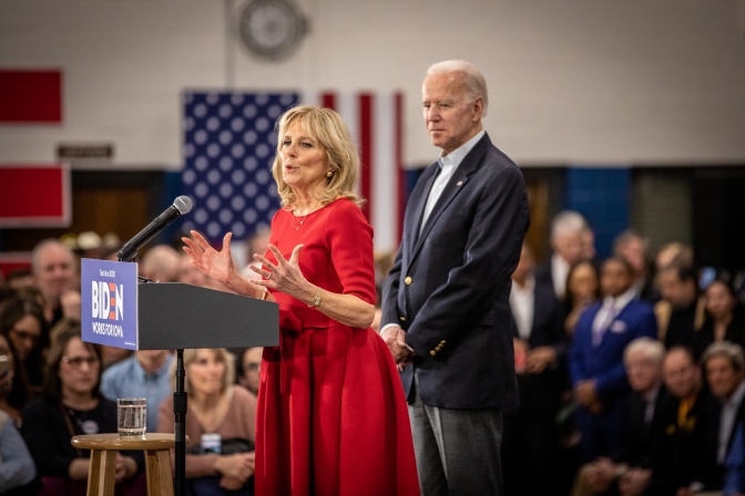 Jill Biden steht in einem roten Kleid an einem Rednerpult und spricht in ein Mikro. Joe Biden steht neben ihr und hört zu. Im Hintergrund eine amerikanische Flagge.