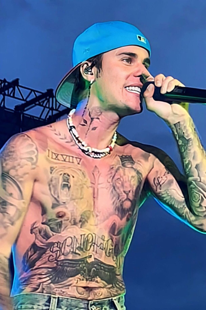 Justin Bieber mit freiem Oberkörper mit vielen Tattoos. Er trägt eine nach hinten gedrehte Schirmmütze und singt in ein Mikrophon.