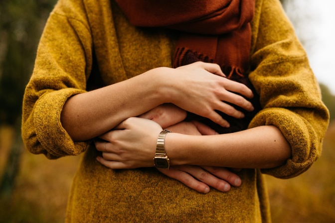Eine Frau in einem senffarbenen Pullover wird von einer anderen Person von hinten umarmt. Man sieht die Gesichter der beiden nicht.
