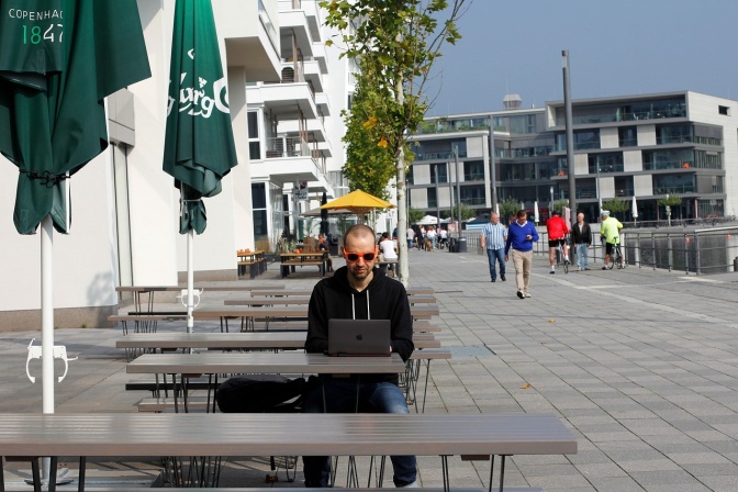 Ein Mann mit Sonnenbrille sitzt in der Fußgängerzone an einem Tisch im Freien. Vor ihm steht ein aufgeklappter Laptop.
