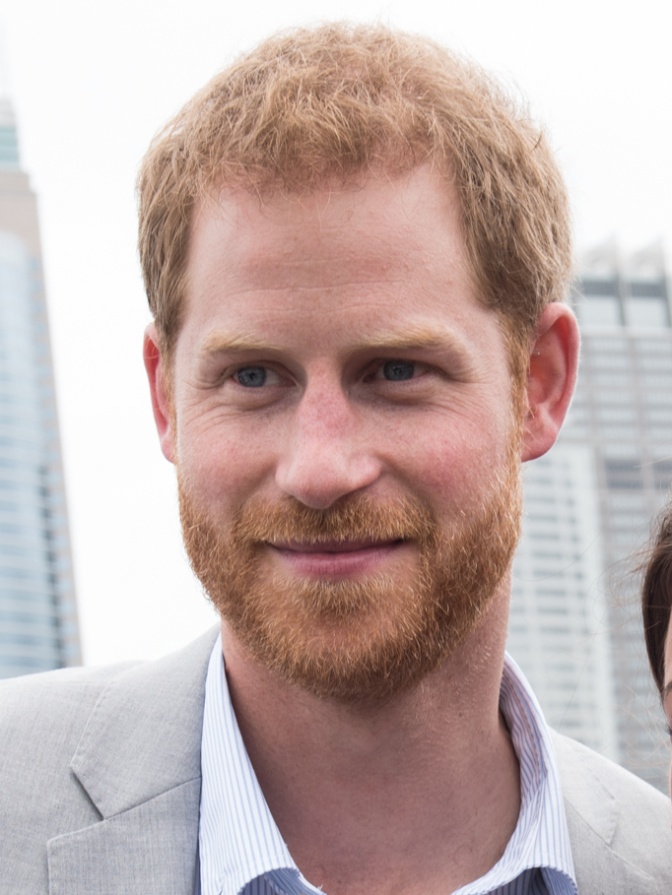 Prinz Harry mit roten Haaren und rotem Bart. Er trägt Hemd und Sakko, im Hintergrund sieht man hohe Gebäude.