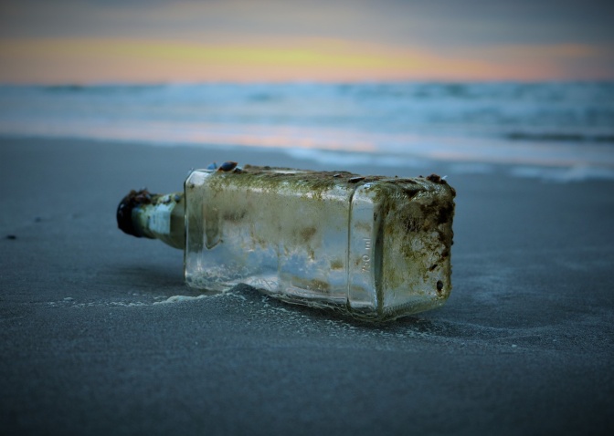 Eine Flaschenpost, zum Teil mit Algen bewachsen, liegt an einem Strand.