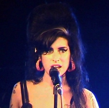 Amy Winehouse mit dunklen, hochgesteckten Haaren und starkem Make up. Sie singt in ein Mikro.