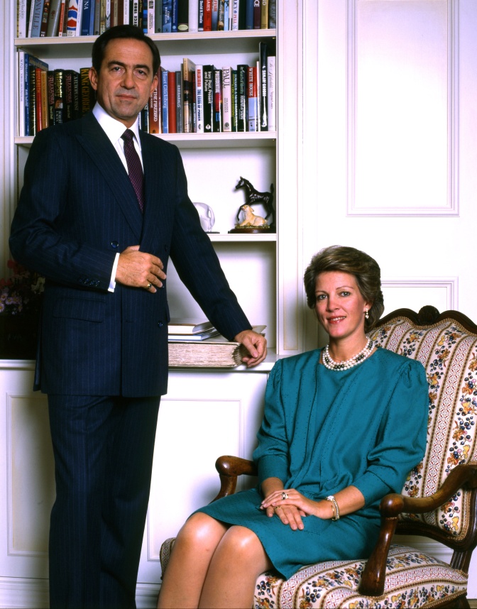 König Konstantin steht neben seiner Frau, die in einem Sessel sitzt. Er trägt Anzug und Krawatte, sie ein petrolfarbenes Kleid.
